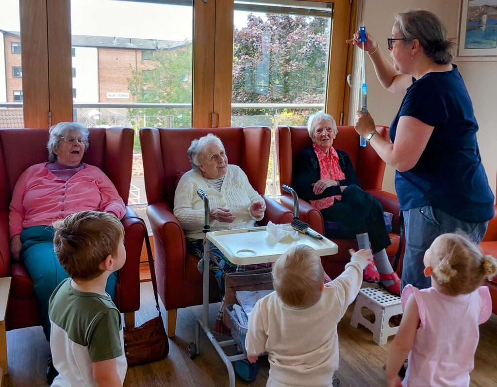 A group of elderly women sit in armchairs while a woman demonstrates something with a syringe to young children standing in front of them in a brightly lit room.