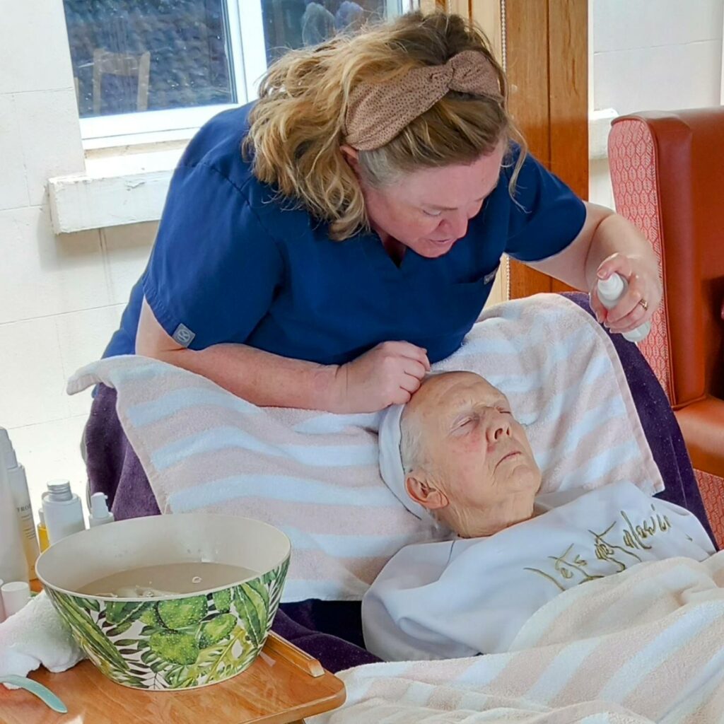 A carer in blue scrubs applies a spray to the forehead of an elderly woman lying back with her eyes closed during a relaxing pamper session facial treatment.