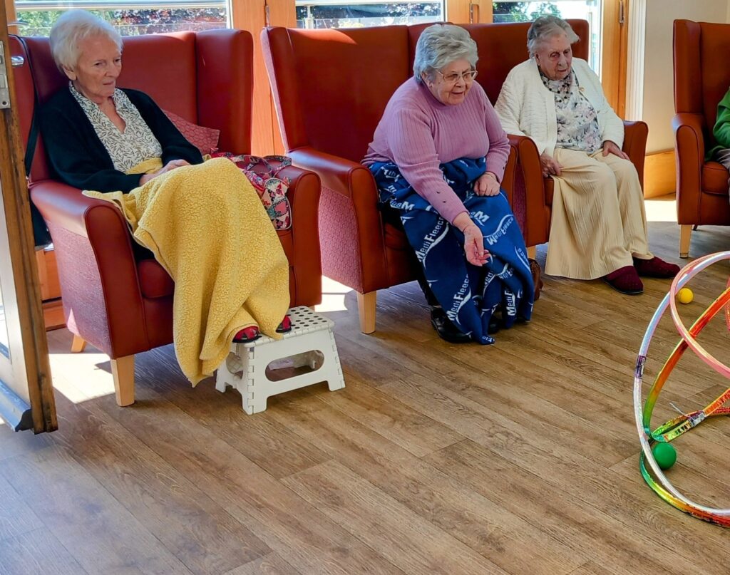 Three elderly women sit in armchairs with blankets over their laps, watching a game of hoop-la on the floor. Broad smiles light up their faces as they cheer each other on.