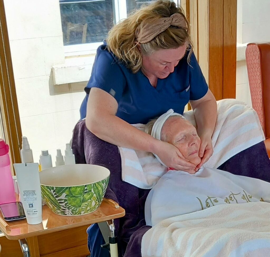 A woman in a blue uniform gives a relaxing facial massage during a pampering session to an elderly person lying back with a towel wrapped around their head in a bright room.