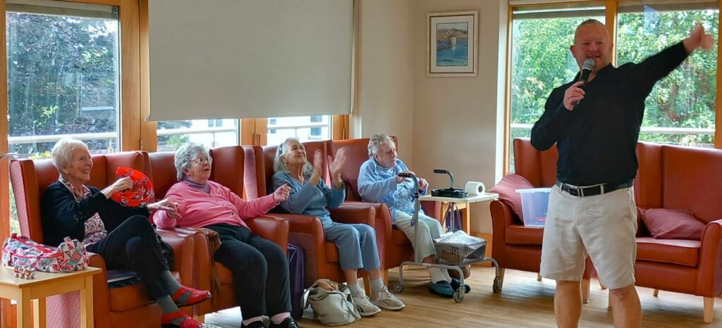 A man holding a microphone sings or speaks to four elderly women seated in armchairs, clapping and smiling, in a well-lit room with large windows.