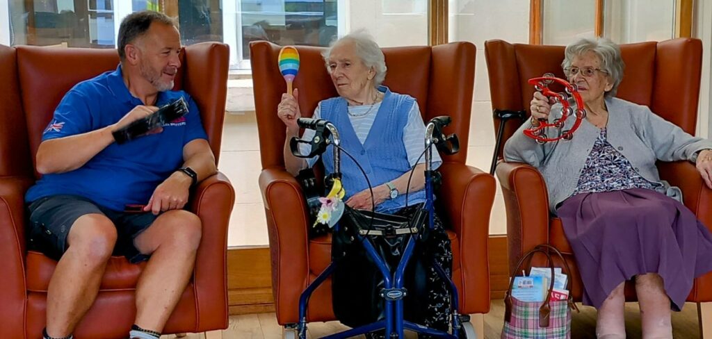Three pensioners sit in armchairs, each holding a musical instrument: a maraca, a tambourine, and a shaker. A walking frame and a handbag are visible beside them.