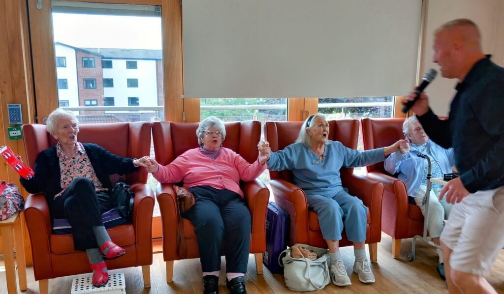 Four elderly women sit in armchairs holding hands while a man with a microphone stands nearby, seemingly entertaining them in a communal room.
