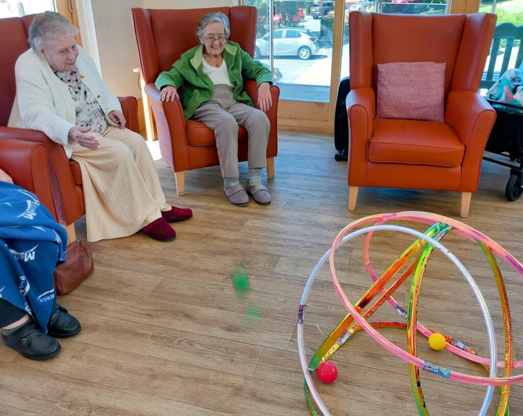 Two elderly women with big smiles sit in armchairs, playing a lively game with colourful hoops and balls on a wooden floor in a bright room.