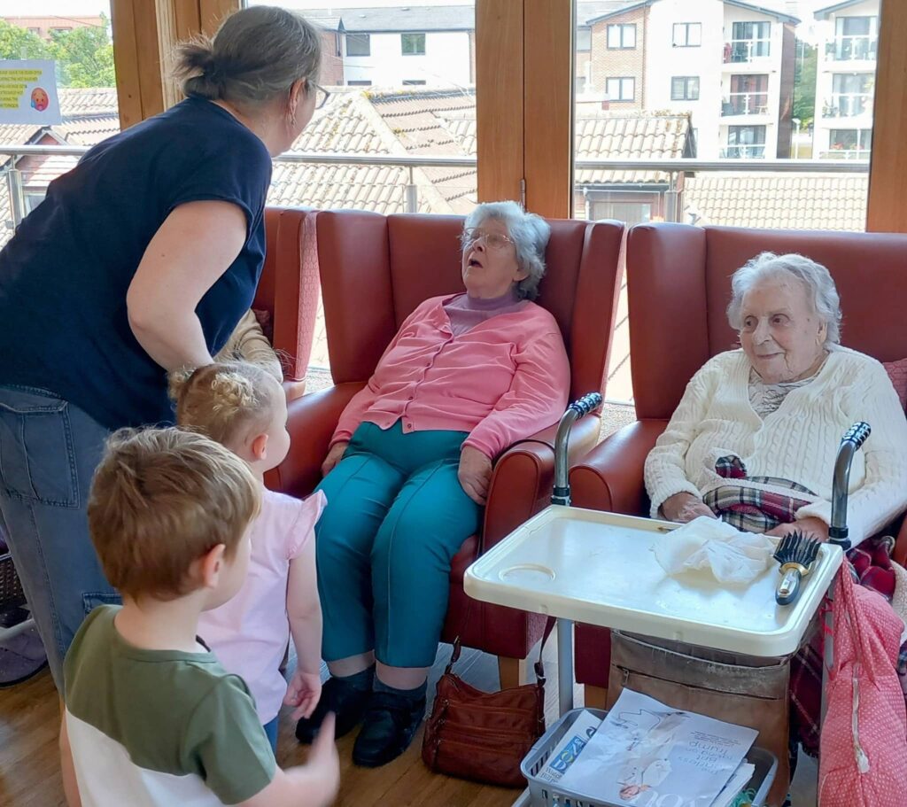 Two elderly women are seated in armchairs by a window; one is dozing while the other is awake. An adult and two children, from Caroline’s Childminding, stand nearby, engaging with them warmly.