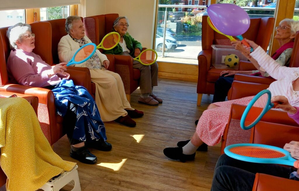 A group of elderly women sit in armchairs in a sunlit room, playing balloon tennis with plastic racquets and a purple balloon, their broad smiles reflecting joy and laughter.