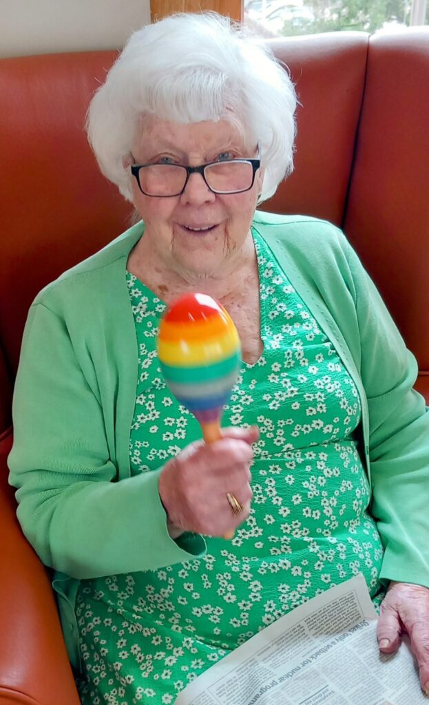 An elderly woman with white hair and glasses is seated on an orange sofa, wearing a green dress and cardigan, holding a colourful maraca, with a newspaper resting on her lap.