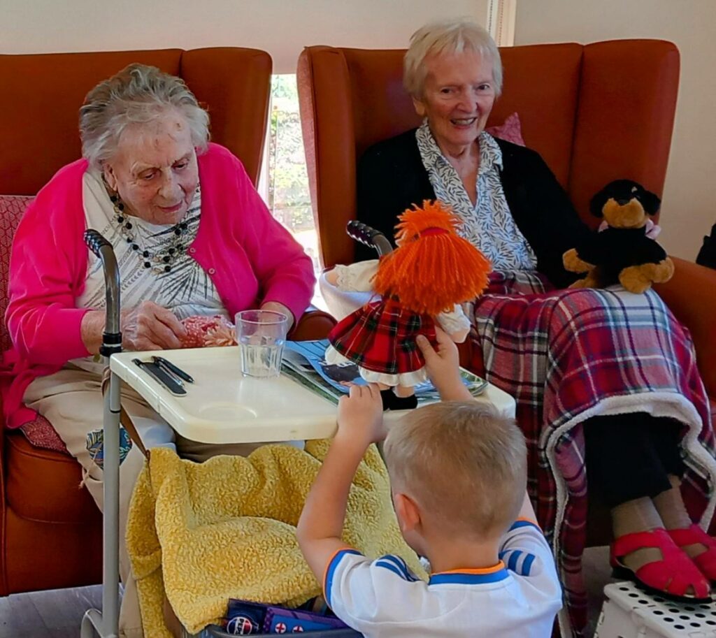 An elderly woman and another older lady sit in armchairs with blankets during a joyful visit, while a young boy from St Agnes Pre-School holds up a doll and soft toy in front of them.