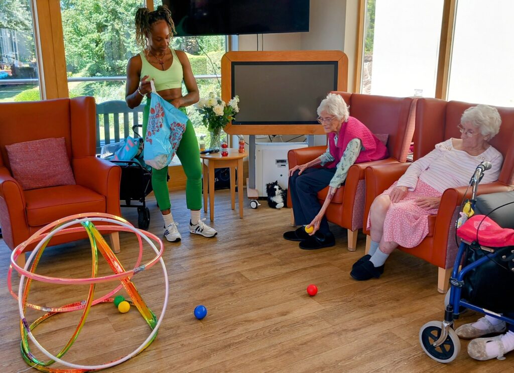 Three elderly women sit in a lounge with big smiles while a woman stands nearby holding a bag of props; a hula hoop structure, colourful balls, and balloon tennis equipment are on the floor.