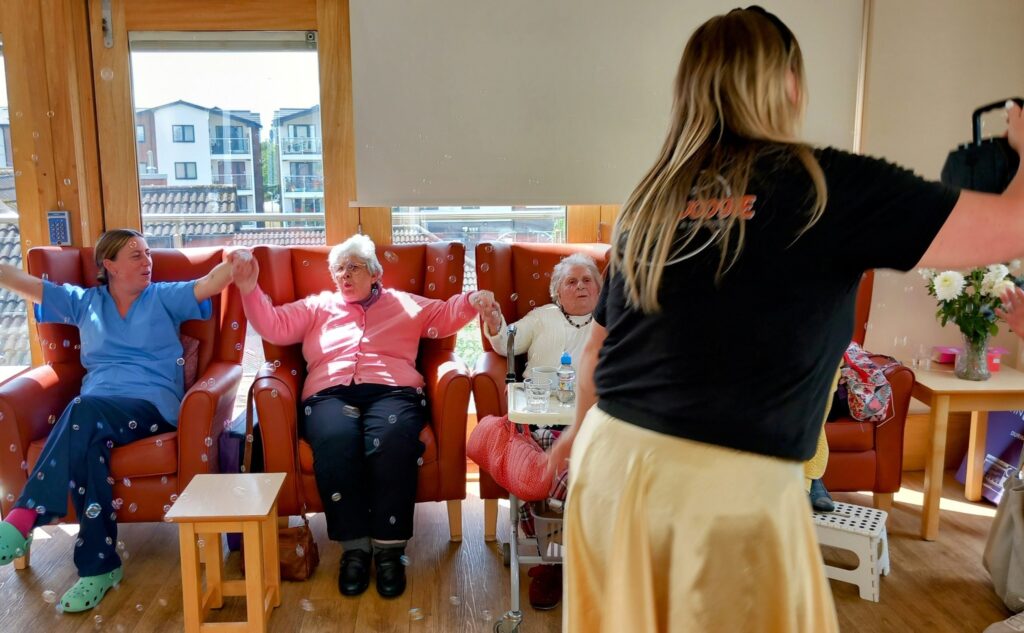 Three elderly women sit in armchairs holding hands while a woman in a black top stands in front of them blowing bubbles in a bright room with large windows.