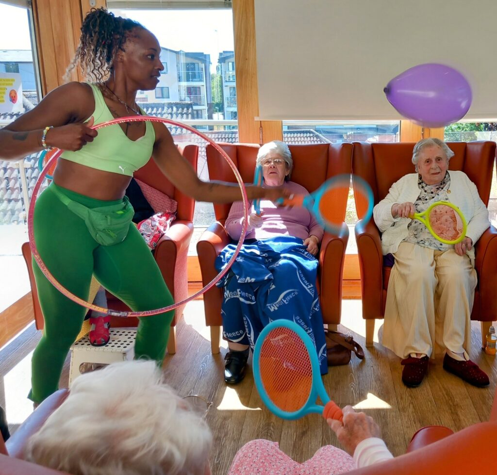 A woman dressed in green leads a group of elderly people in balloon tennis, using rackets, a balloon, and a hula hoop. The group sits on chairs, sharing broad smiles as they take part in this enjoyable indoor activity.
