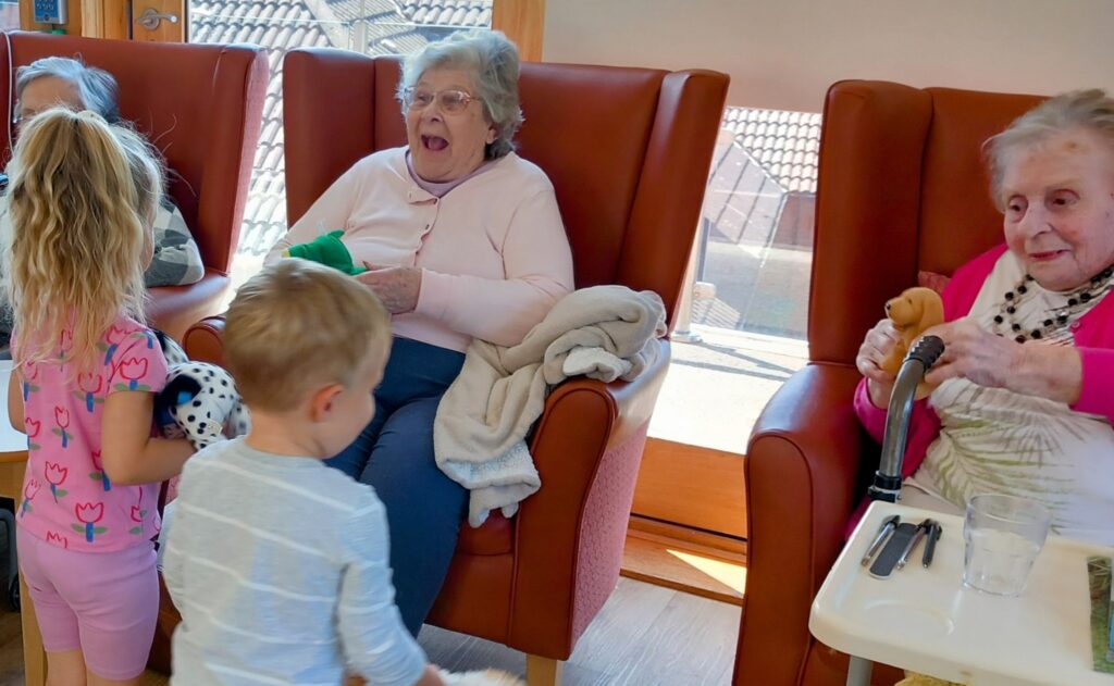 During a joyful visit to St Agnes Pre-School, two young children interact with three elderly women seated in chairs; one woman is smiling and holding a green toy, whilst another holds a small stuffed dog.