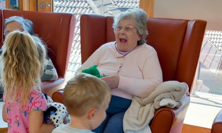 During a joyful visit to St Agnes Pre-School, two young children interact with three elderly women seated in chairs; one woman is smiling and holding a green toy, whilst another holds a small stuffed dog.