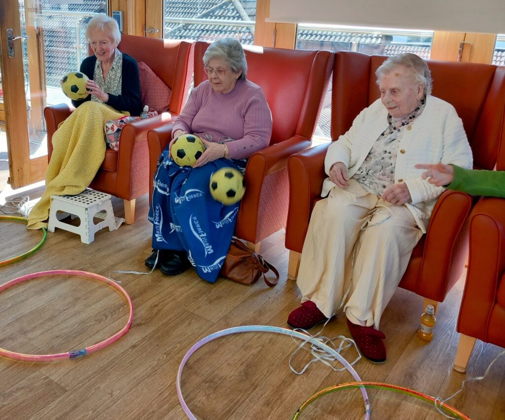 Three elderly women sit in armchairs with big smiles, holding yellow and black footballs, while colourful hula hoops are placed on the floor in front of them inside a bright room.