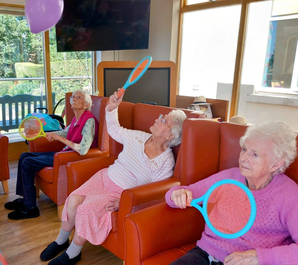 Three elderly women with broad smiles, seated in armchairs, play balloon tennis indoors using plastic racquets to hit balloons as part of a lively group activity.