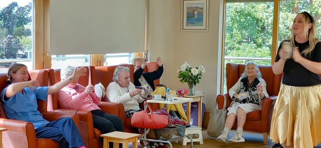 A group of elderly women and a carer sit in a lounge, clapping and moving their arms, while a woman stands at the front of the room leading an activity.
