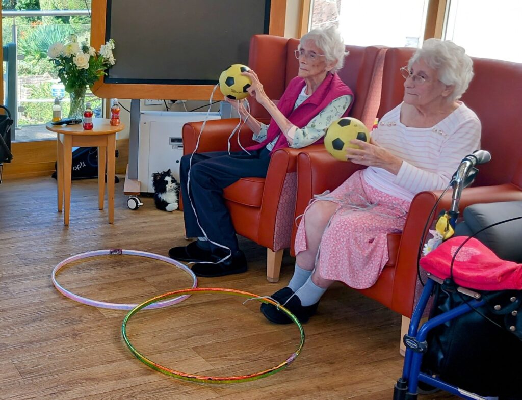 Two elderly women with broad smiles sit in armchairs, holding yellow footballs attached to strings, with hula hoops on the floor in front of them in a well-lit room.