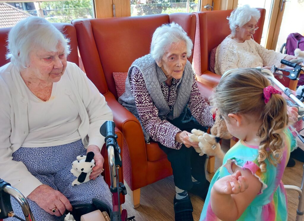Three elderly women sit in armchairs, enjoying a delightful visit as a young girl from St Agnes Pre-School shows them a soft toy in the cosy care home setting.