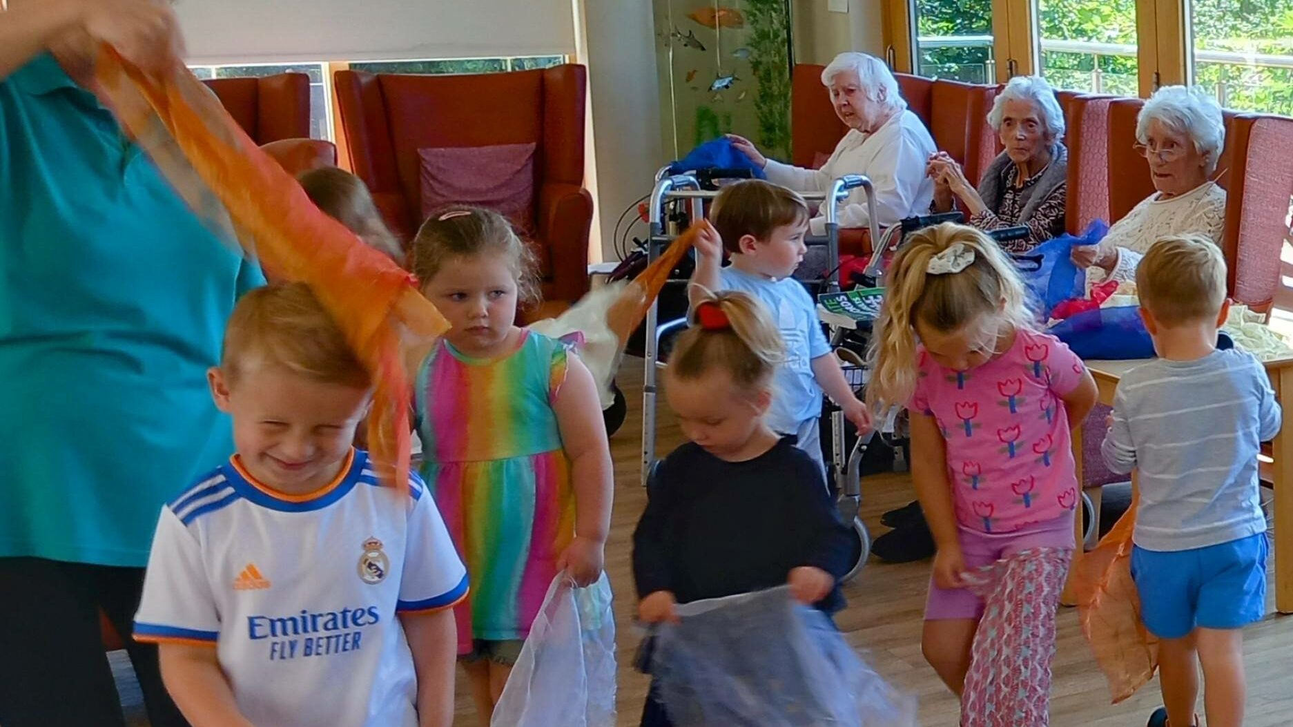 Young children from St Agnes Pre-School, holding coloured scarves, walk in a line at a care home, bringing a joyful visit to elderly residents seated and watching in the background.