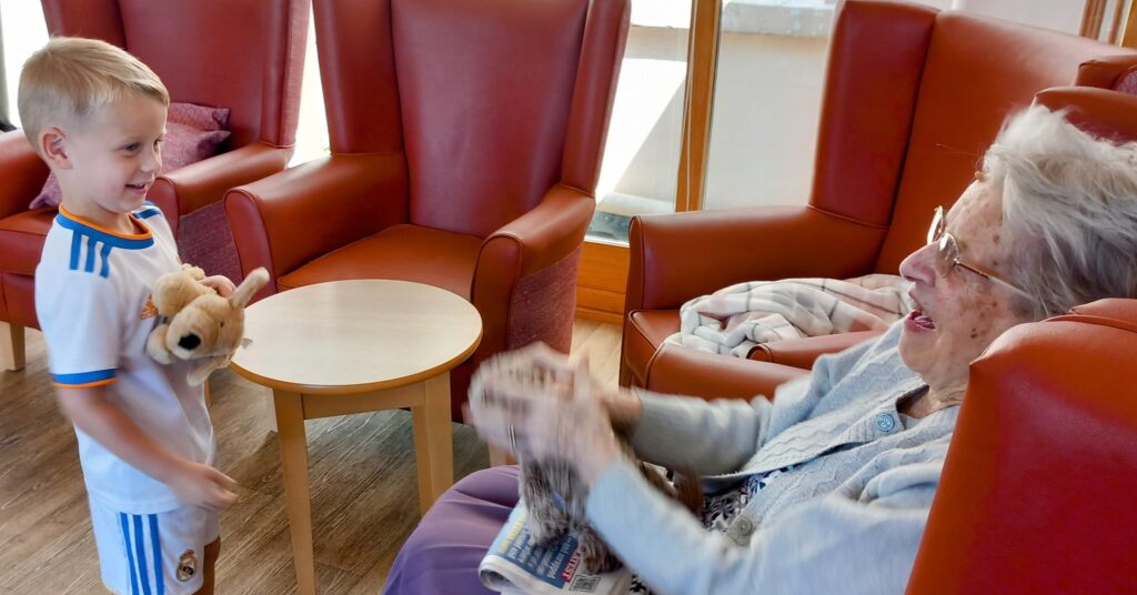 A young boy from St Agnes Pre-School holding a soft toy smiles at an elderly woman who is clapping her hands; both seem to be enjoying a delightful visit together.