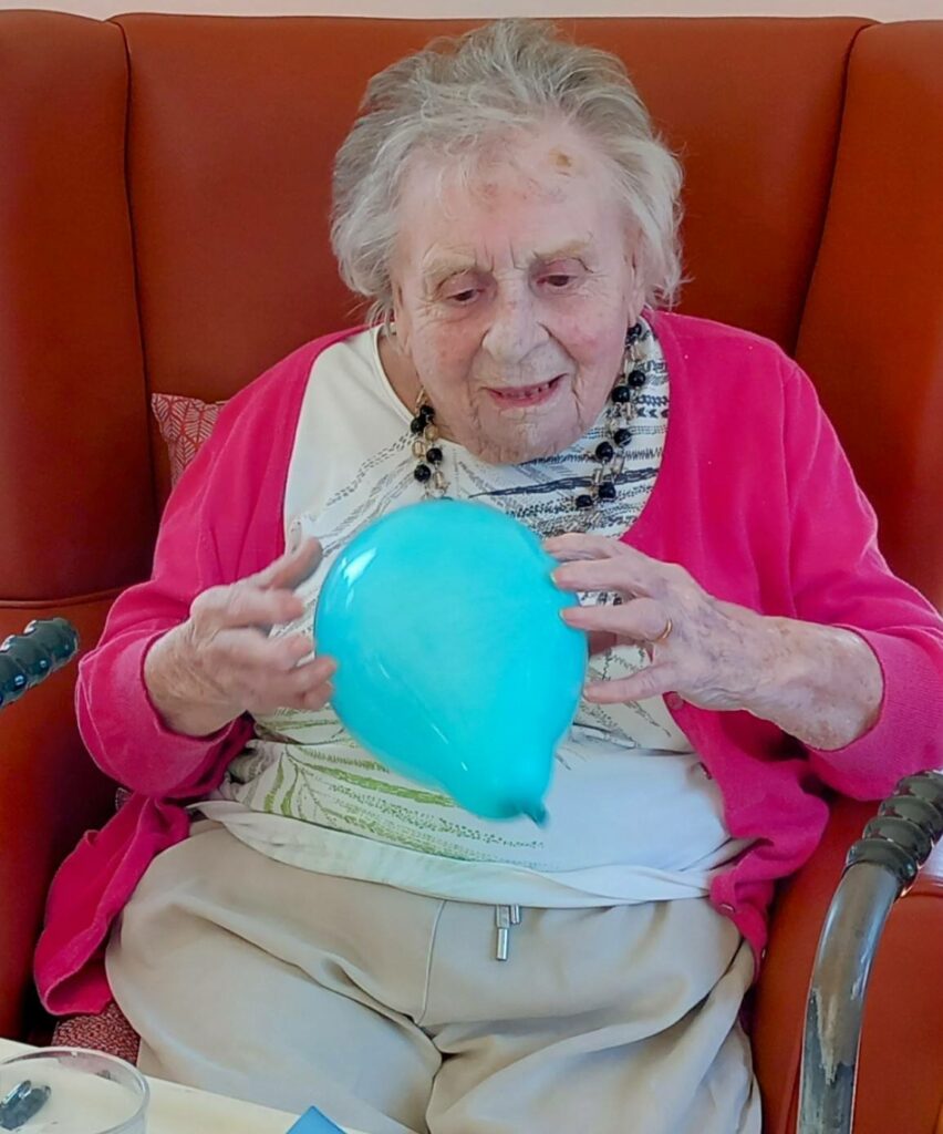 An elderly woman sits in an armchair holding a blue balloon with both hands, her pink cardigan and beige trousers reflecting the warmth of a joyful visit from St Agnes Pre-School.