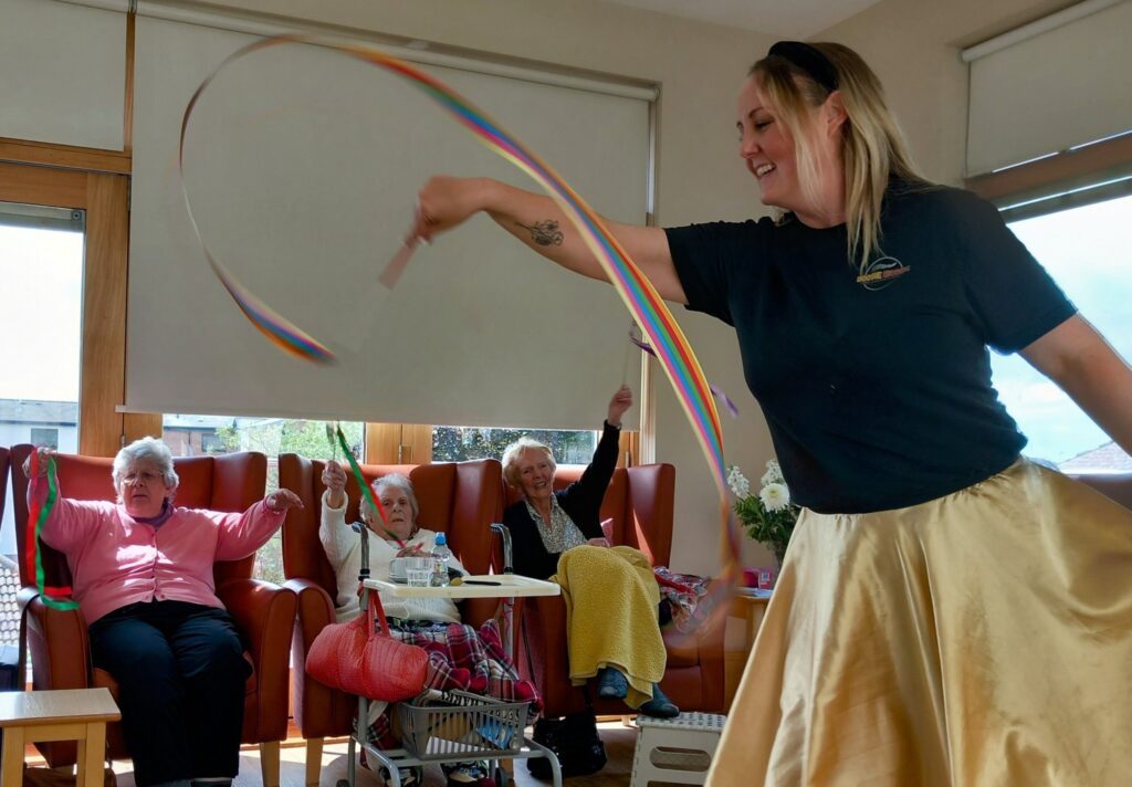 A woman waves a colourful ribbon while three elderly women, seated in armchairs, watch and join in with their own ribbons in a bright room.