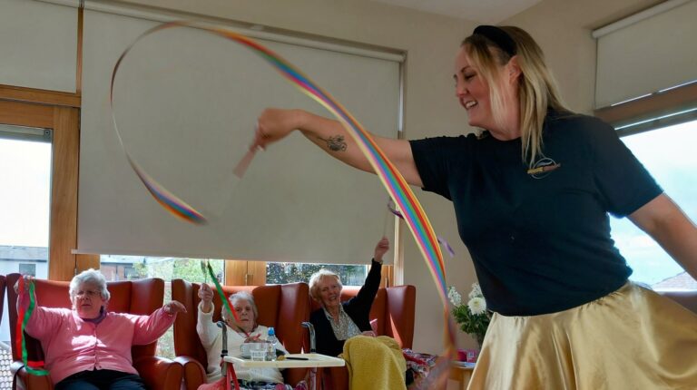 A woman waves a colourful ribbon while three elderly women, seated in armchairs, watch and join in with their own ribbons in a bright room.