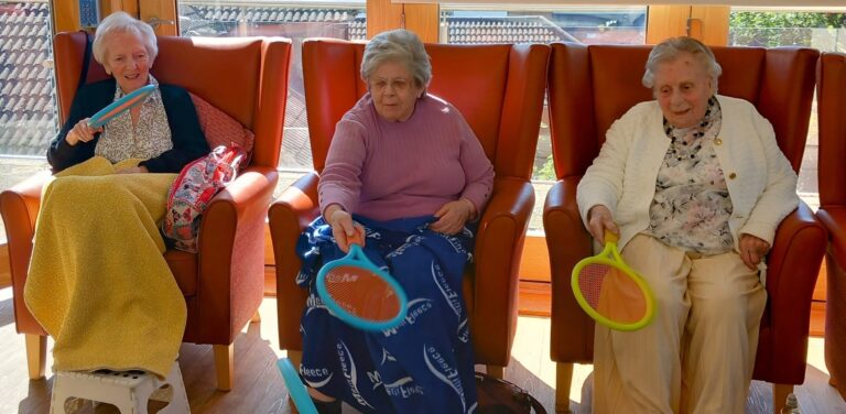 Three elderly women with broad smiles sit in armchairs, each holding a plastic bat and enjoying a lively game of balloon tennis indoors.
