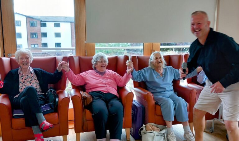 Three elderly women are seated in armchairs, holding hands and smiling, while a man with a microphone stands nearby, also smiling.