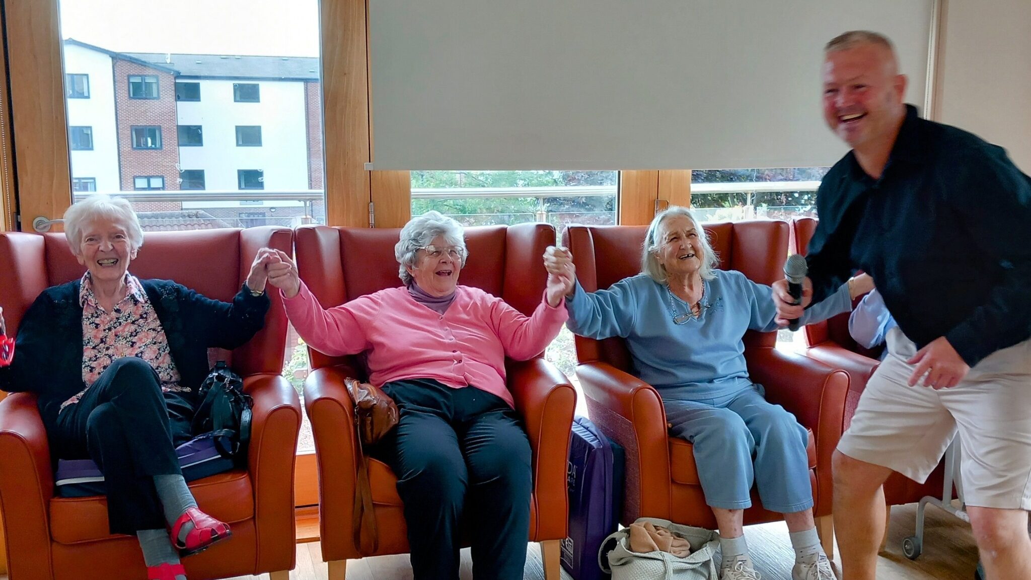 Three elderly women are seated in armchairs, holding hands and smiling, while a man with a microphone stands nearby, also smiling.