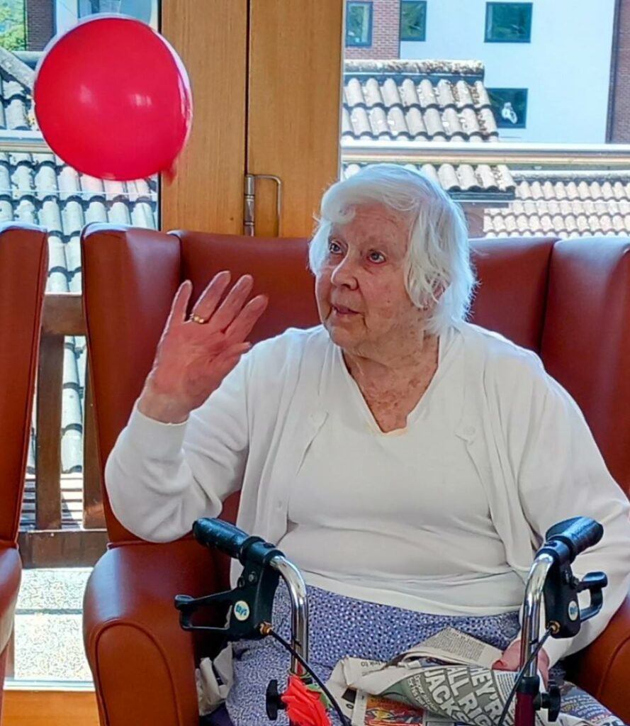 Elderly woman sits in an armchair with a Zimmer frame, raising her hand towards a floating red balloon during a joyful visit, with a newspaper on her lap.