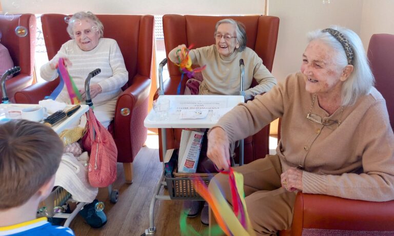 Three elderly women, seated in chairs, smile and wave colourful ribbons as they interact with a young child during the final session of Popcats at St Agnes Pre-School in a brightly lit room.