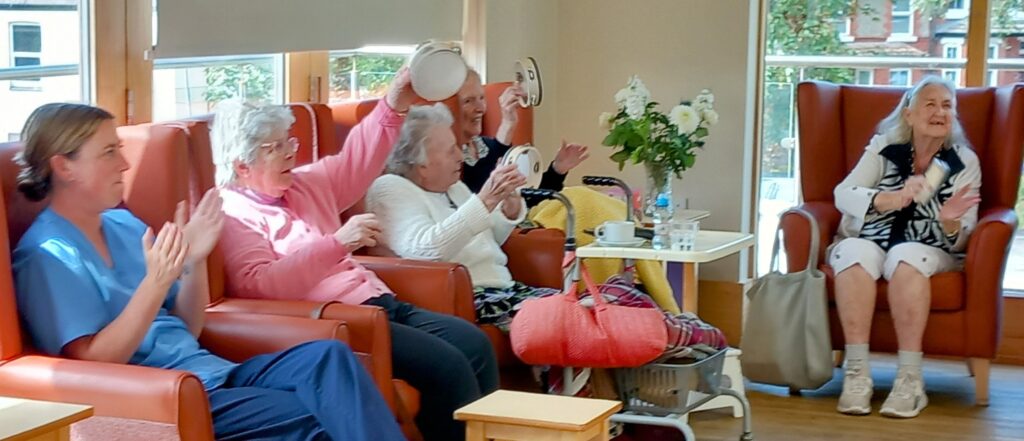 Five elderly women are seated in a bright room; some are clapping while others hold up tambourines, appearing to be taking part in an activity together.