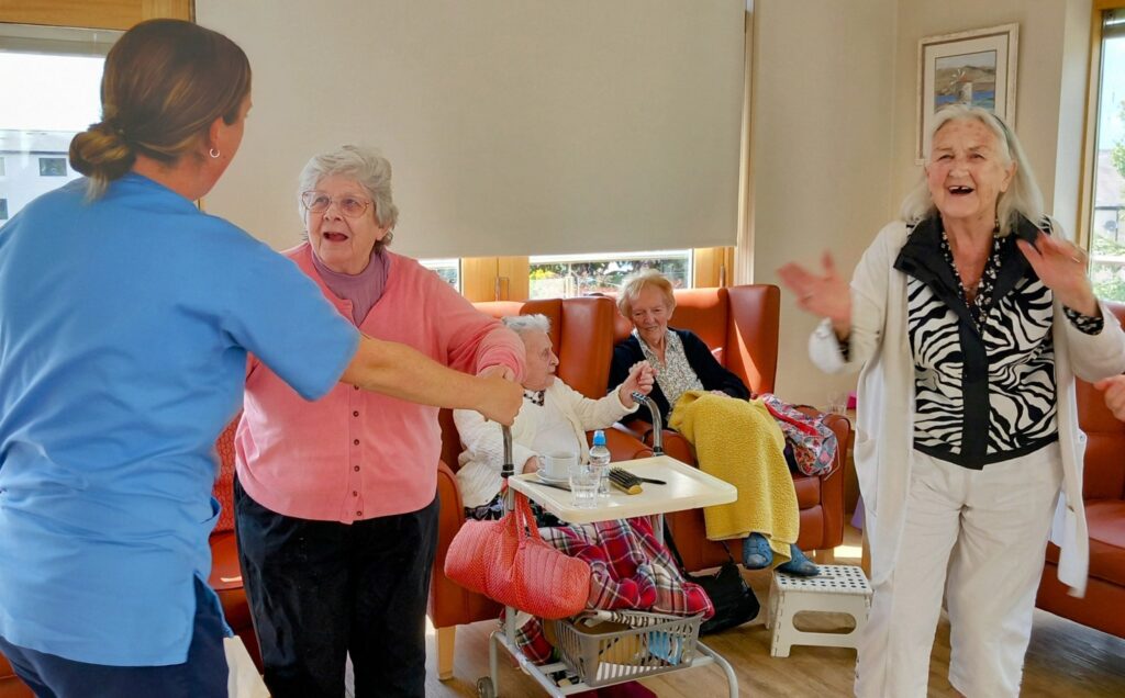 A group of elderly women, some seated and some standing, are happily interacting with a carer in a bright, comfortable room.