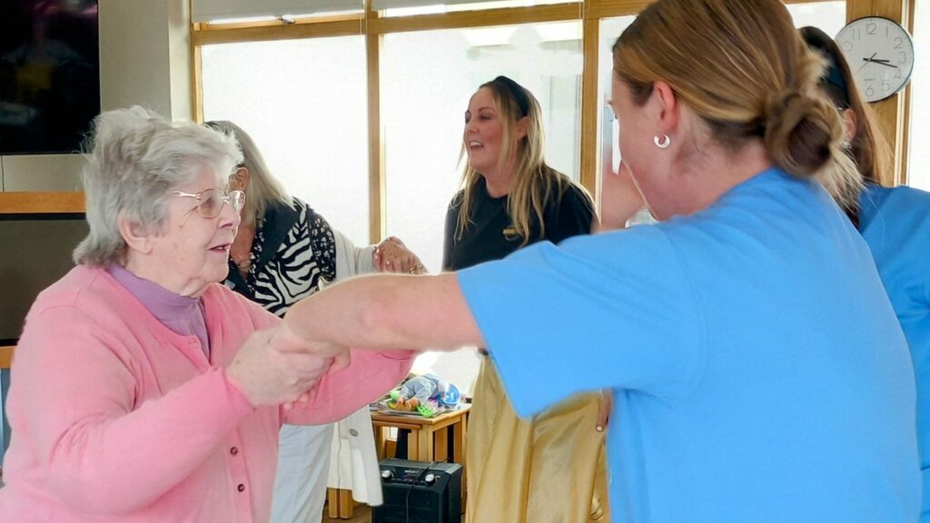 An elderly woman in a pink jumper dances with a carer in blue scrubs, while two other women smile in the background of a bright room.