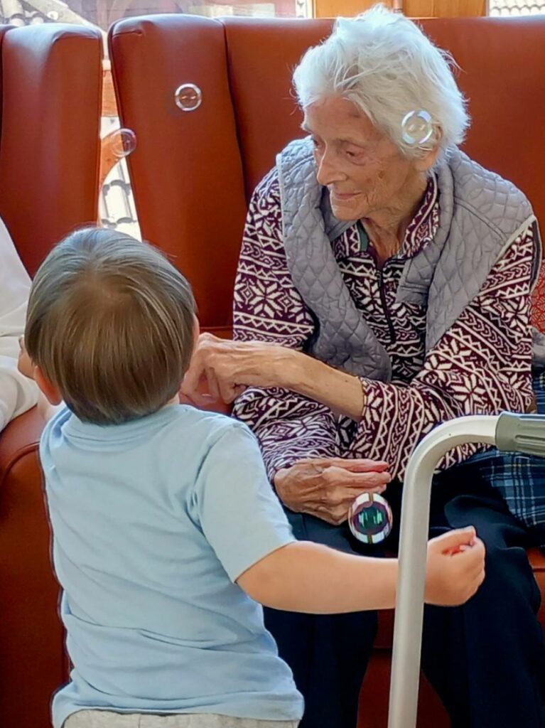 An elderly woman sits in a chair, enjoying a joyful visit with a young child blowing bubbles, while a walking frame stands nearby—capturing the warmth of connection at St Agnes Pre-School.