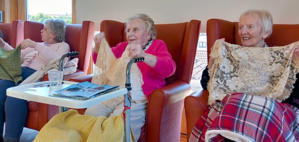 Three elderly women sit in orange chairs, holding lace fabric; one has a walking frame, and a blanket covers their laps. Balloons hint at a joyful visit, with water and magazines on the tray in front of them.