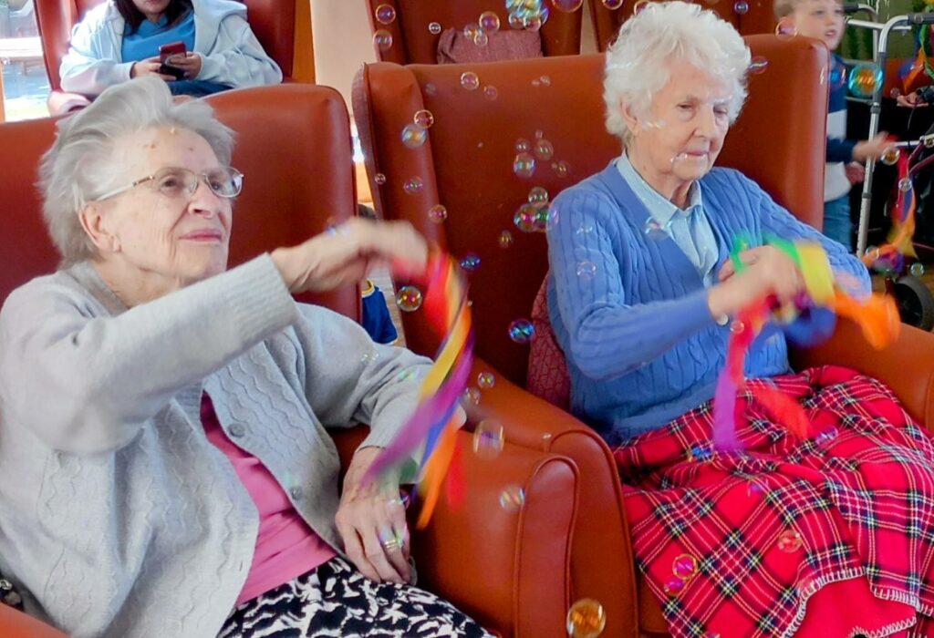 Two elderly women seated in chairs wave colourful ribbons at a Joyful Send-Off, with bubbles floating around them. Other people can be seen in the background during this lively Popcats session.