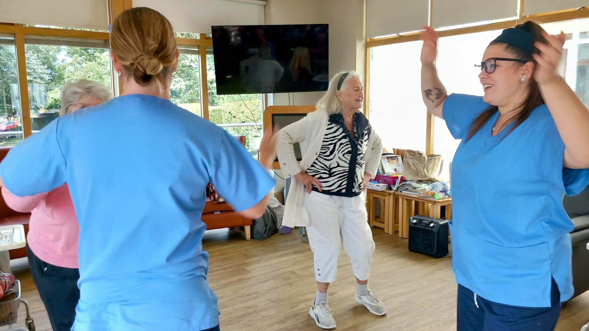Two healthcare workers and two elderly women are standing and dancing together in a bright room with large windows and a television.