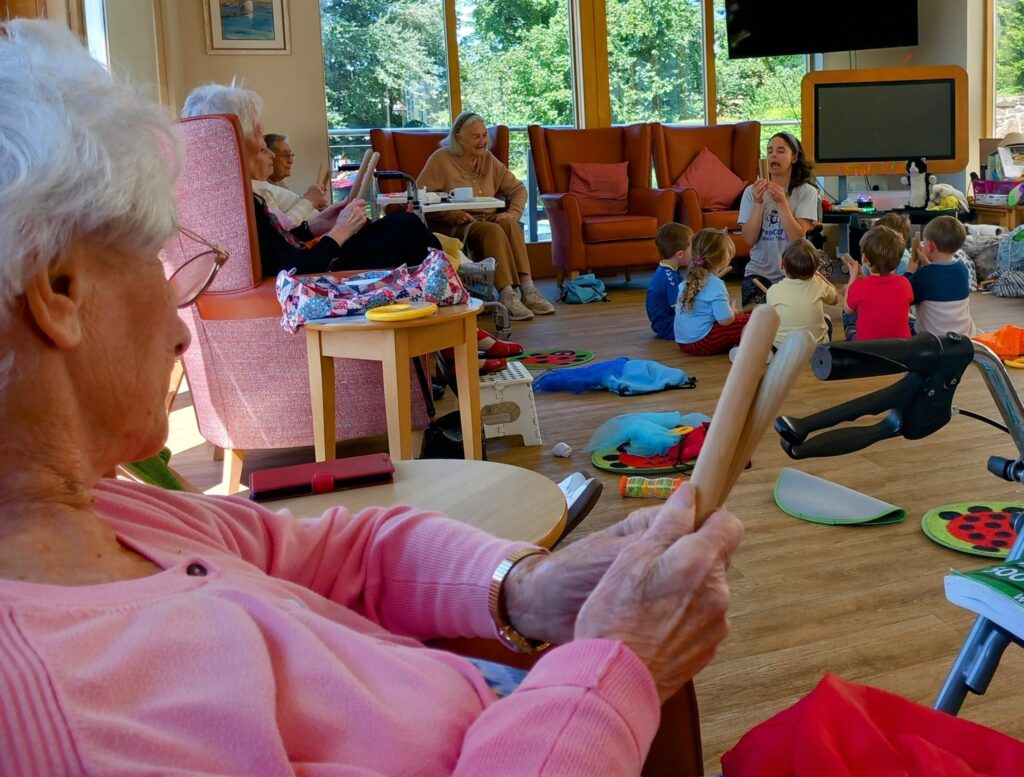 Older adults and young children from St Agnes Pre-School take part in the final session of Popcats in a bright room, with a woman holding a wooden object in the foreground.