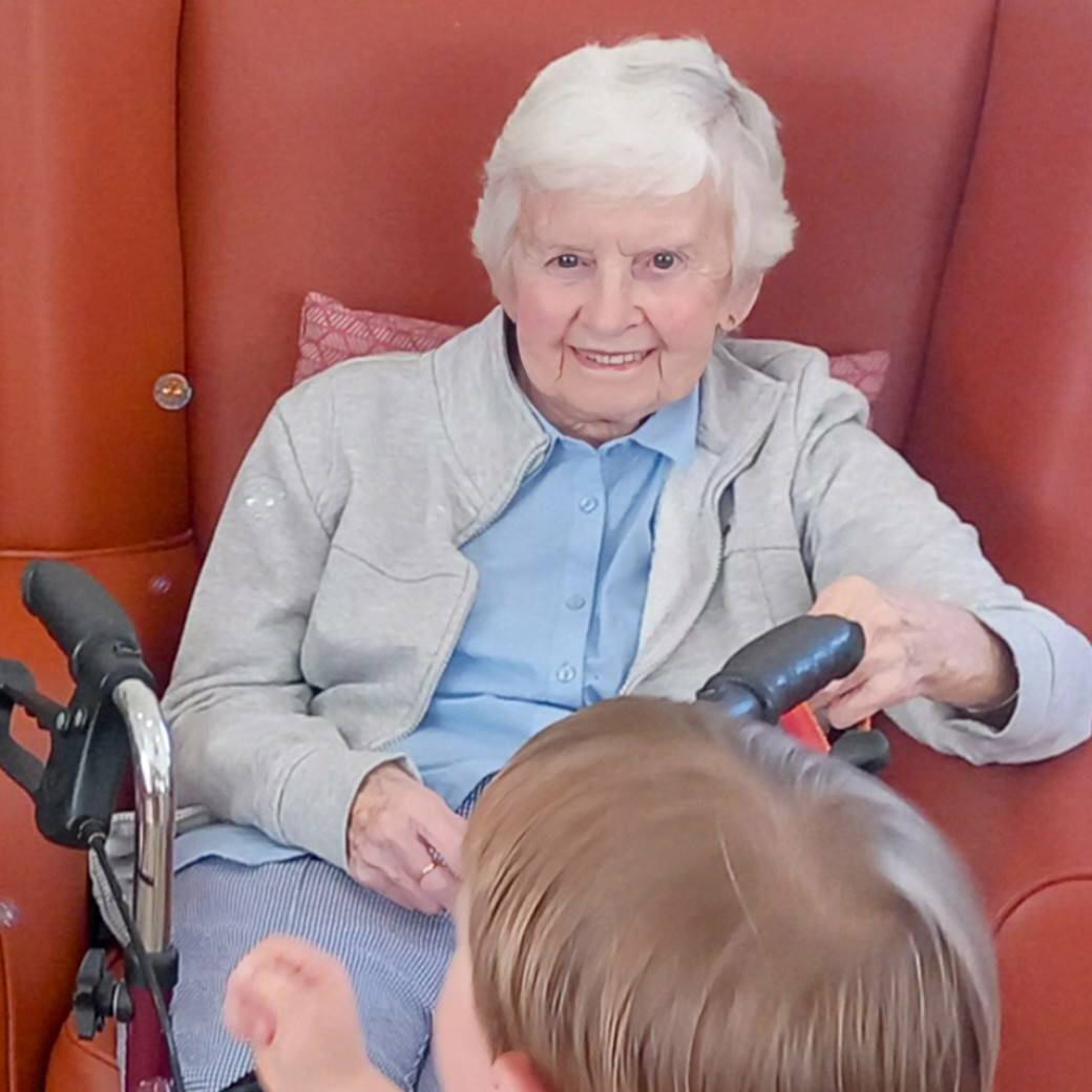 An elderly woman sits in a chair with a walking frame in front of her, smiling at a young child from St Agnes Pre-School during the final session.