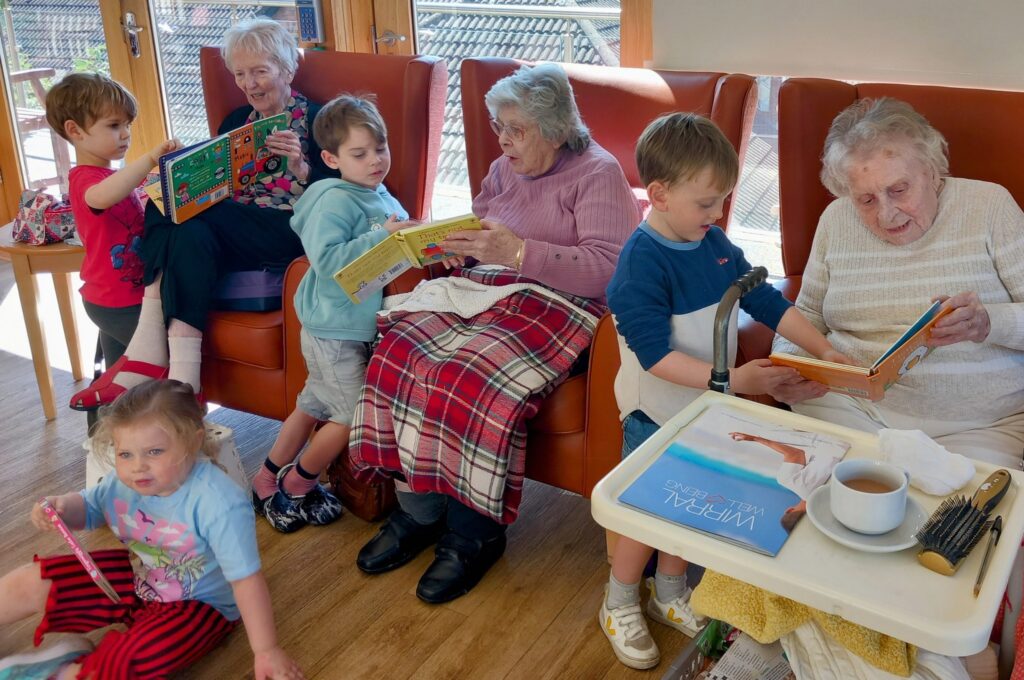 Children and elderly women sit together in a well-lit room at St Agnes Pre-School, reading books; tea and a magazine rest on a tray beside one of the older women. (British English version:) Children and elderly women are seated together in a well-lit room at St Agnes Pre-School, reading books; tea and a magazine sit on a tray beside one of the older women.