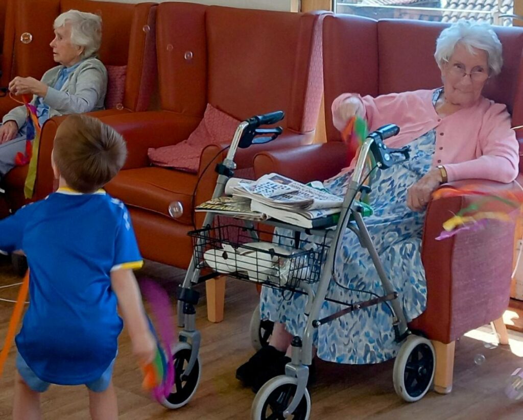 An elderly woman sits in a chair with a walking frame, holding colourful ribbons during a joyful farewell. A young boy and another elderly woman play with ribbons nearby at St Agnes Pre-School.