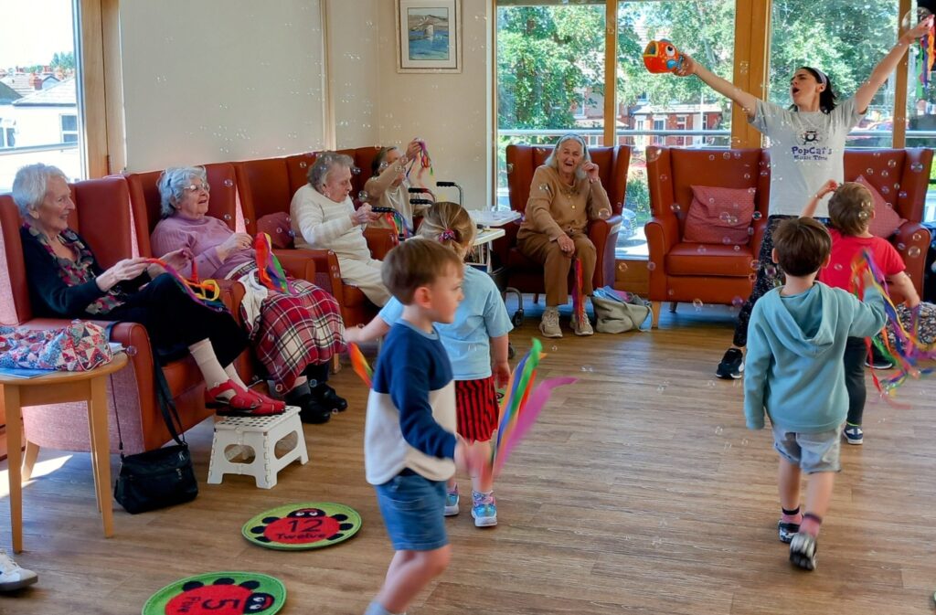 Children from St Agnes Pre-School play with ribbon wands and bubbles during their final session, as elderly women look on and smile in a brightly lit communal room with large windows and orange chairs.