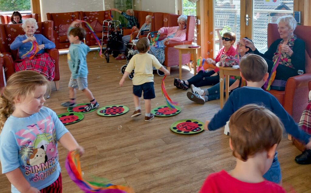 Children play with colourful ribbon wands and toys on the floor while elderly people watch and smile in a bright, sunlit room during the final session of Popcats at St Agnes Pre-School.