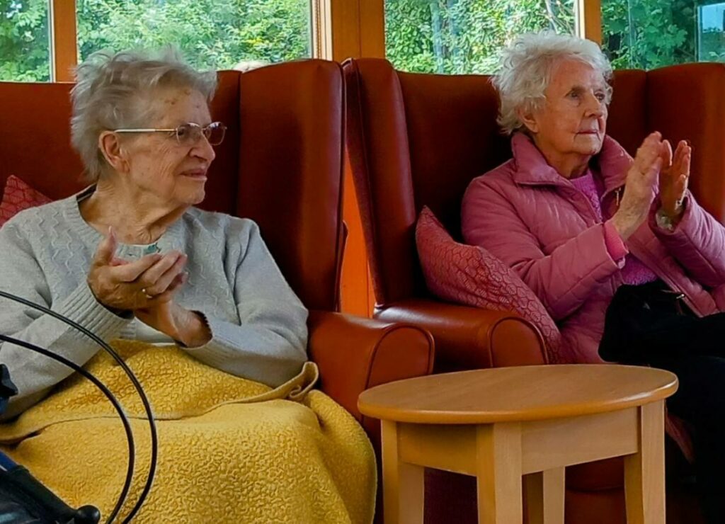 Two elderly women sit indoors on red chairs, applauding. One is wearing a grey jumper and a yellow blanket; the other has on a pink jacket. Green foliage is visible through the window behind them.