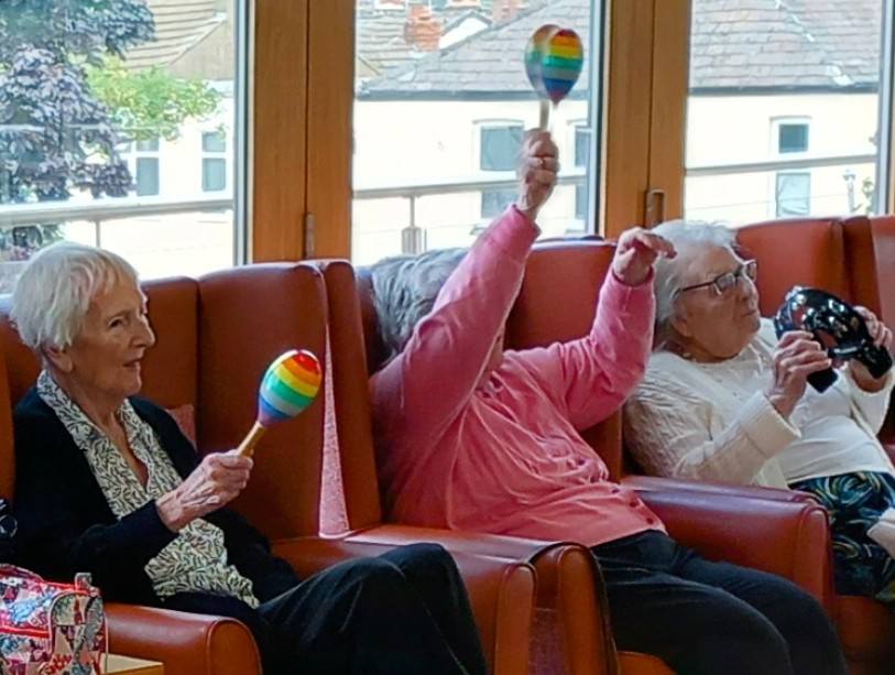 Three elderly women are seated in armchairs, holding colourful musical instruments, with one woman raising a rainbow maraca.
