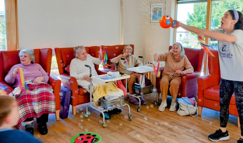 A group of elderly women sitting in armchairs reach out to touch bubbles during a joyful Popcats session, while a woman stands using a bubble blower in a bright room.