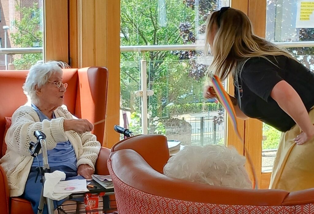 An elderly woman sitting in a chair extends her hand towards a standing woman holding a rainbow ribbon in a brightly lit room with large windows.