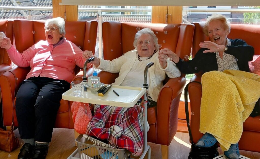 Three elderly women sit in armchairs, holding hands, smiling and laughing. One has a walking frame and a tray with water and other items. Sunlight streams in through the window behind them.